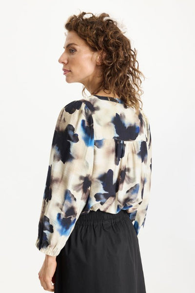 Kvinne i bluse med blått og svart blomstertrykk, knapper foran, lange ermer med elastiske mansjetter.