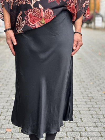 Person iført svart satengskjørt med skinnende overflate og subtil vidde nederst, på brosteinsvei.