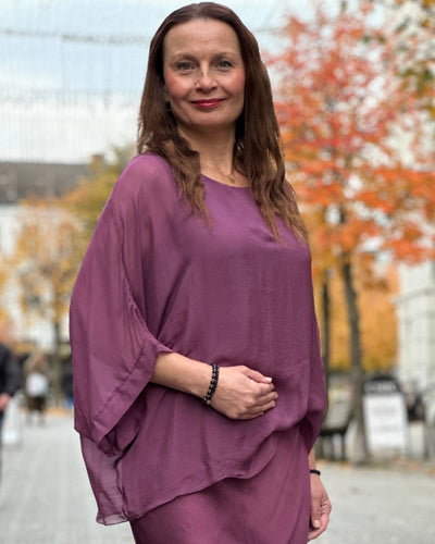 Kvinne i lilla to-lags bluse med gjennomsiktig ytterlag, vide ermer og rund hals.
