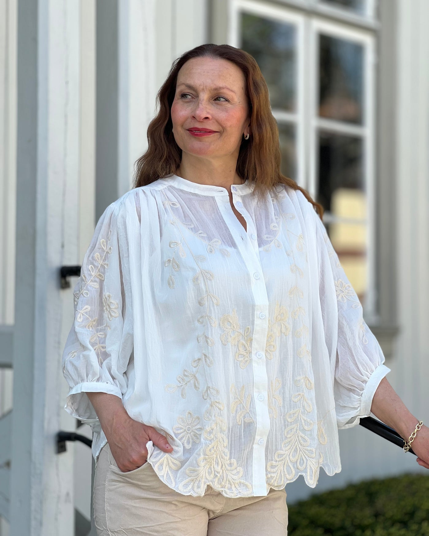 Kvinne i hvit chiffonbluse med broderte blomster, står foran et vindu utendørs.