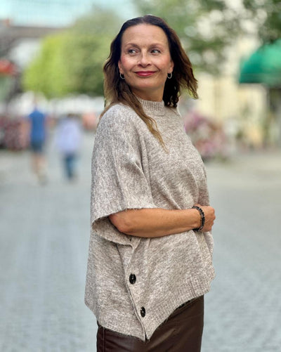 Modell med lys brun poncho med høy hals og knapper, i et uskarpt utendørs miljø.