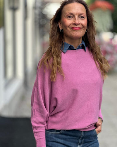 Kvinne i lys rosa genser med løs passform, brede ermer, høy hals, og denimskjorte under.