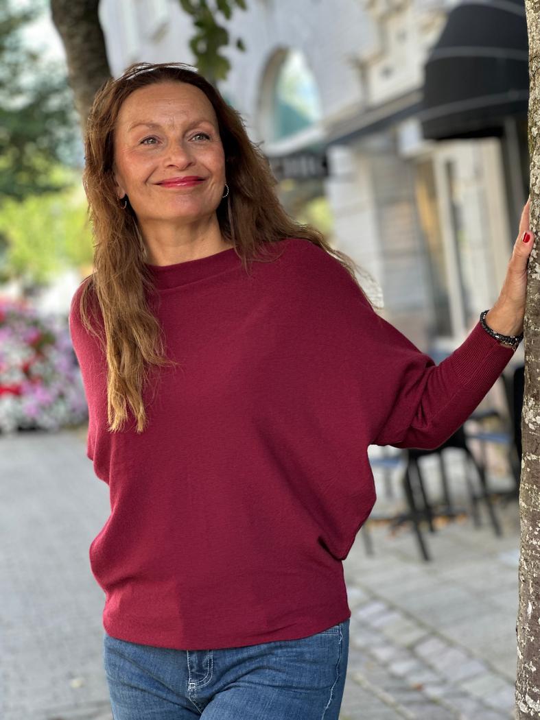 Kvinne i dyp rød batgenser, står ved trestamme, smiler. Genseren har løs passform og lange ermer.