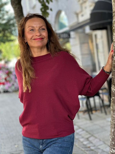 Kvinne i dyp rød batgenser, står ved trestamme, smiler. Genseren har løs passform og lange ermer.