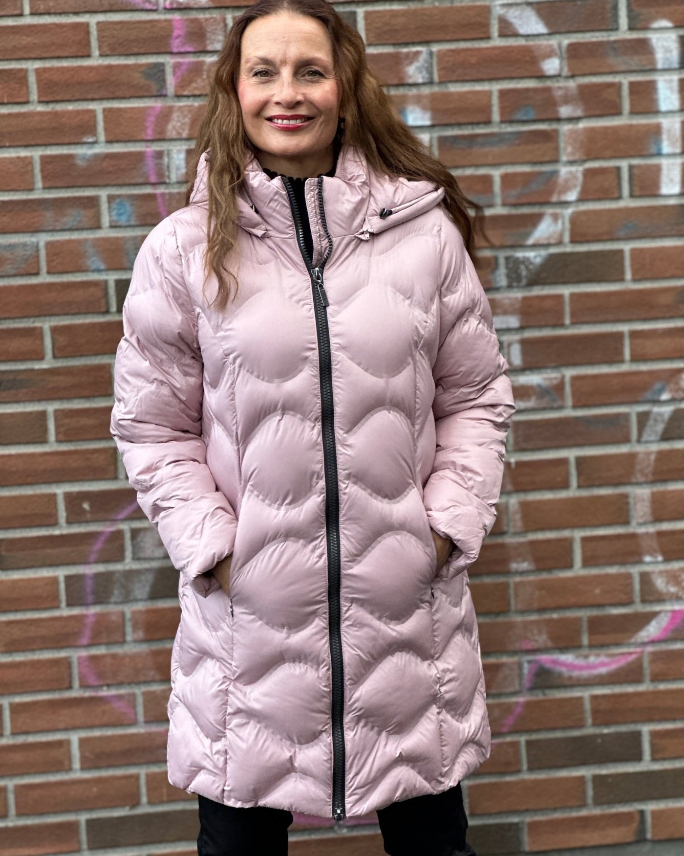 Rosa quiltet jakke med hette og glidelås, kvinne foran murvegg med graffiti.