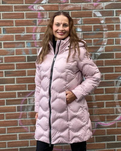 En kvinne i rosa quiltet jakke med hette og glidelås, står foran en murvegg med graffiti.