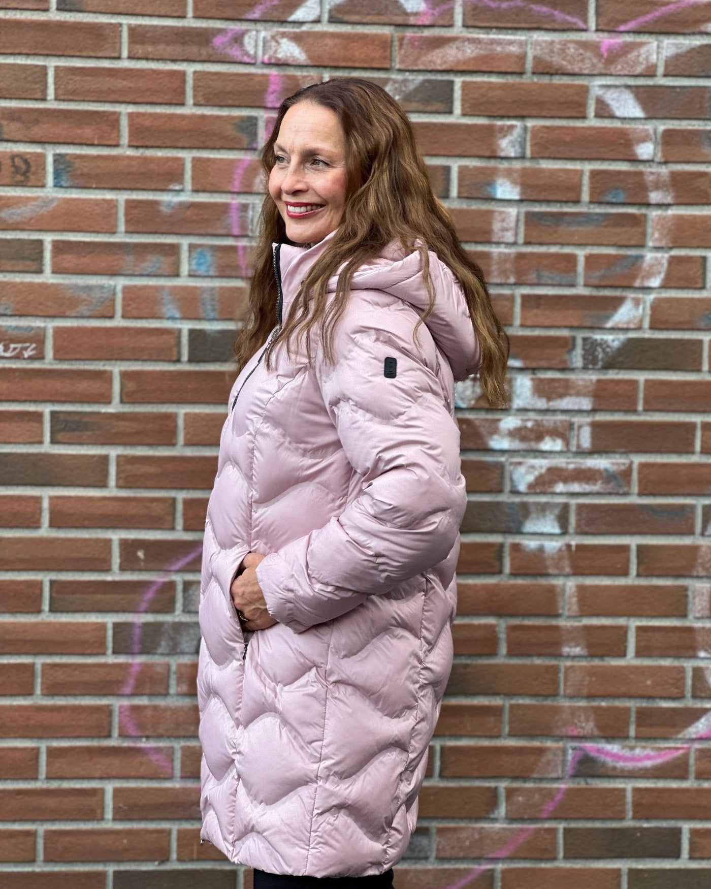 Kvinne i rosa, quiltet jakke med hette, står i profil mot en murvegg.