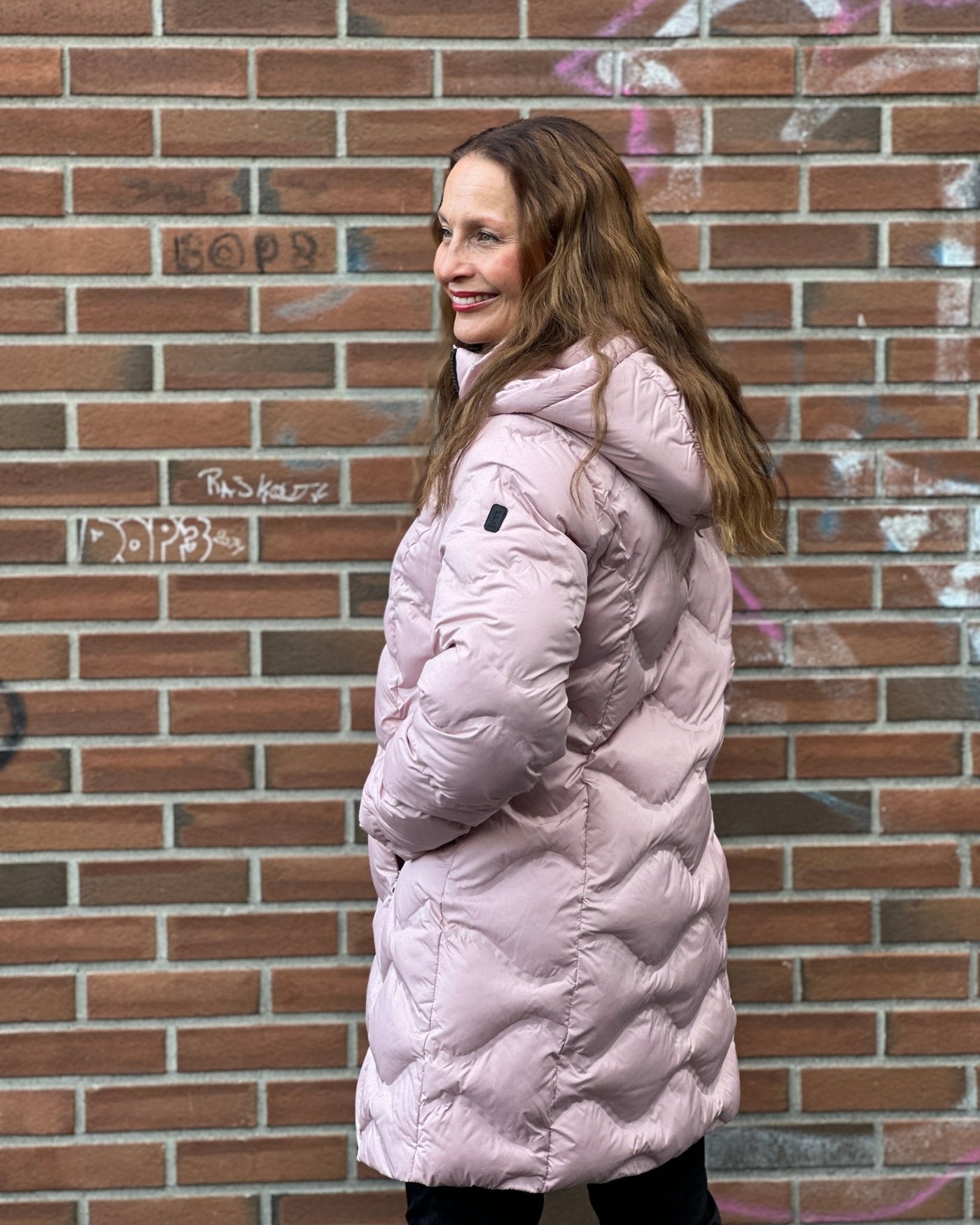 Kvinne i rosa quiltet jakke med hette, står mot murvegg, smiler og ser til siden.