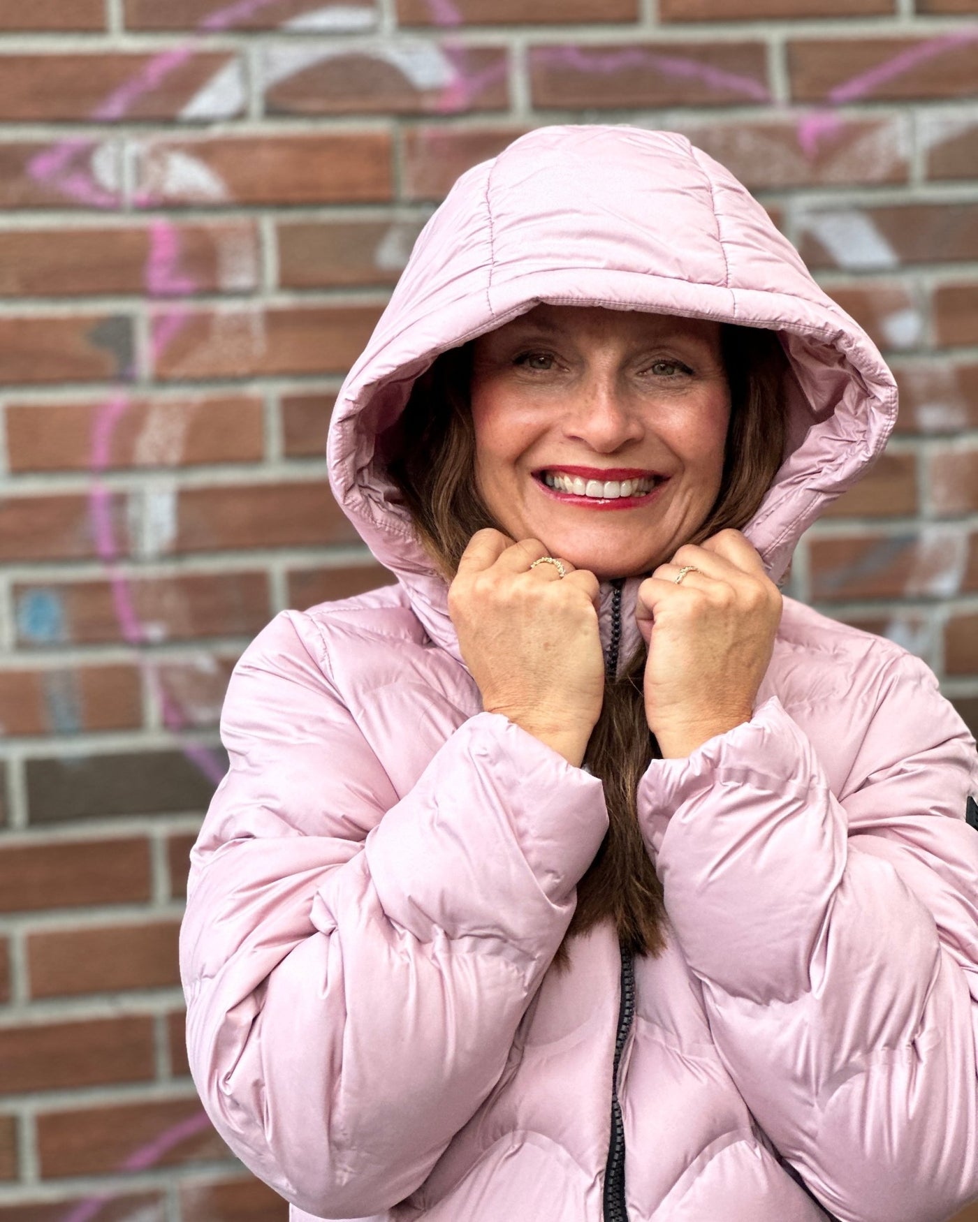 Kvinne i rosa quiltet jakke med hette, smilende foran en teglsteinsvegg.