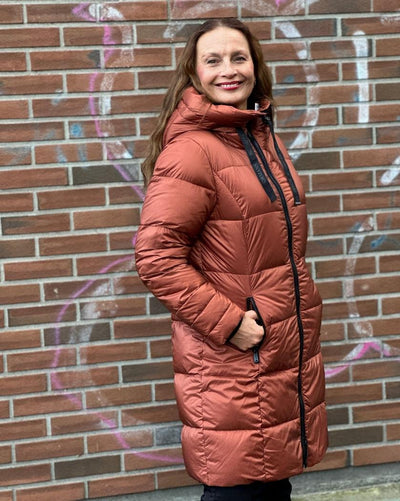 Kvinne i lang, rustfarget dunjakke med hette og glidelås, mot en murvegg med graffiti.