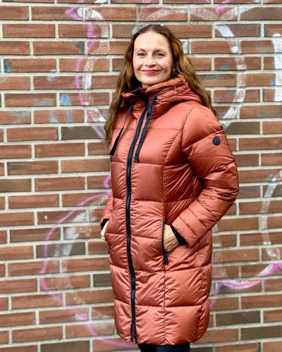 Kvinne i kobberfarget quiltet dunjakke med hette og snorer, foran glidelås, mot murvegg.