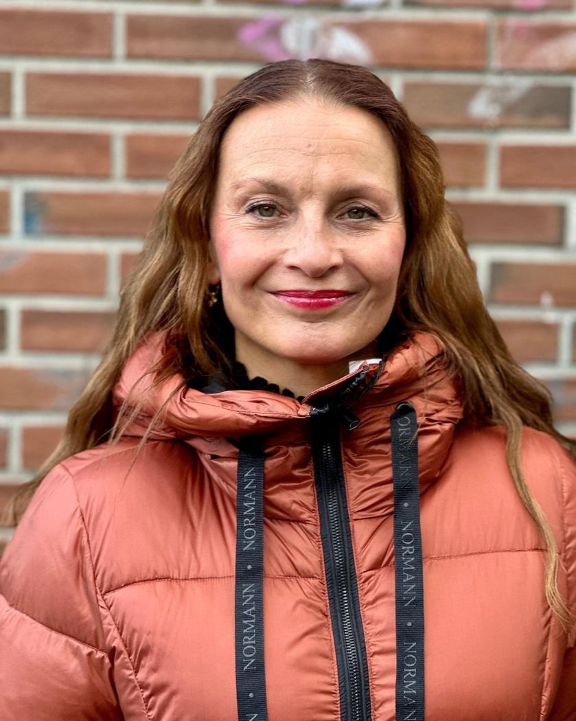 Person med kobberfarget, quiltet dunjakke, hette med "NORMANN" snorer, foran glidelås, murvegg bak.