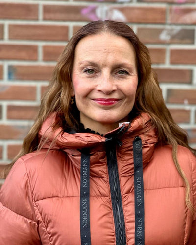 Person med kobberfarget, quiltet dunjakke, hette med "NORMANN" snorer, foran glidelås, murvegg bak.