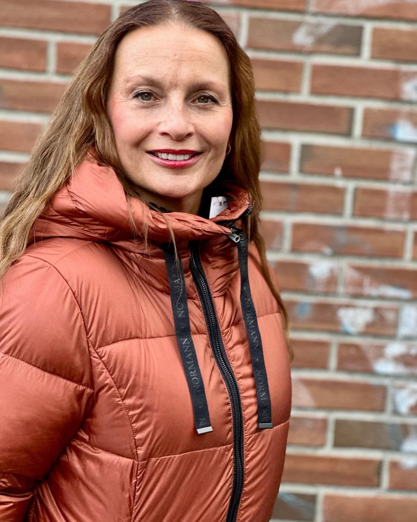Kvinne med oransje quiltet jakke med hette og snorer mot en murvegg.
