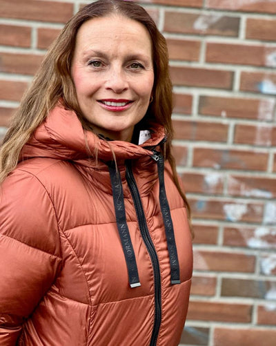 Kvinne med oransje quiltet jakke med hette og snorer mot en murvegg.