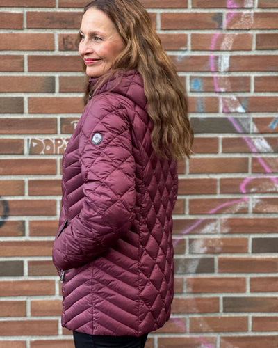 Person iført burgunder quiltet jakke med hette og glidelåslommer, mot en murvegg med graffiti.