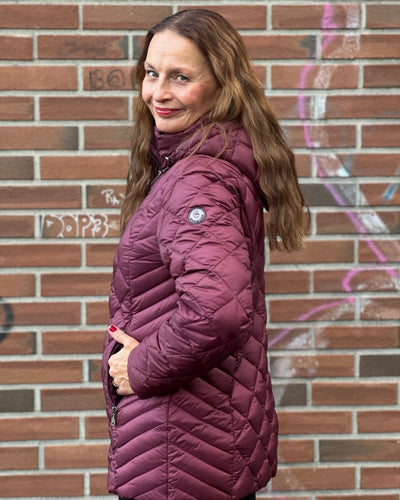 Person i lilla quiltet jakke med hette, glidelåslommer og logo på ermet, foran en graffitivegg.