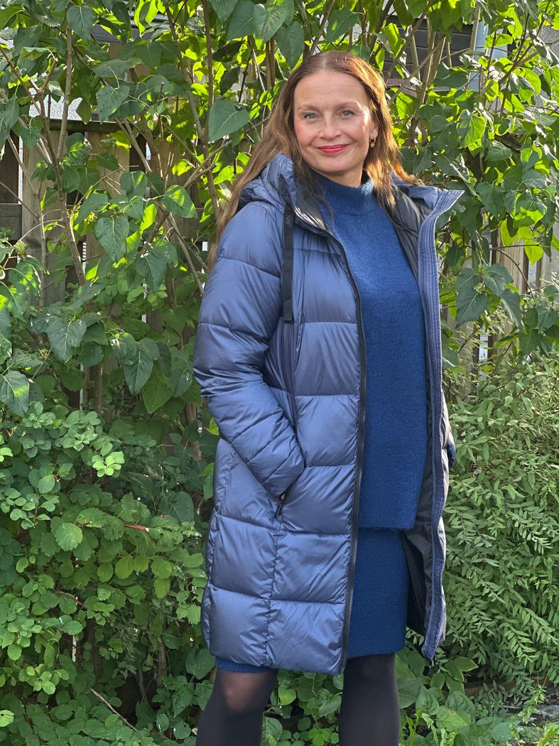 Kvinne i lang, blå dunjakke med hette og glidelås, står foran grønne busker utendørs.