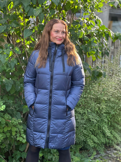 Kvinne i blå quiltet dunjakke med hette og glidelås, står foran grønn bakgrunn utendørs.