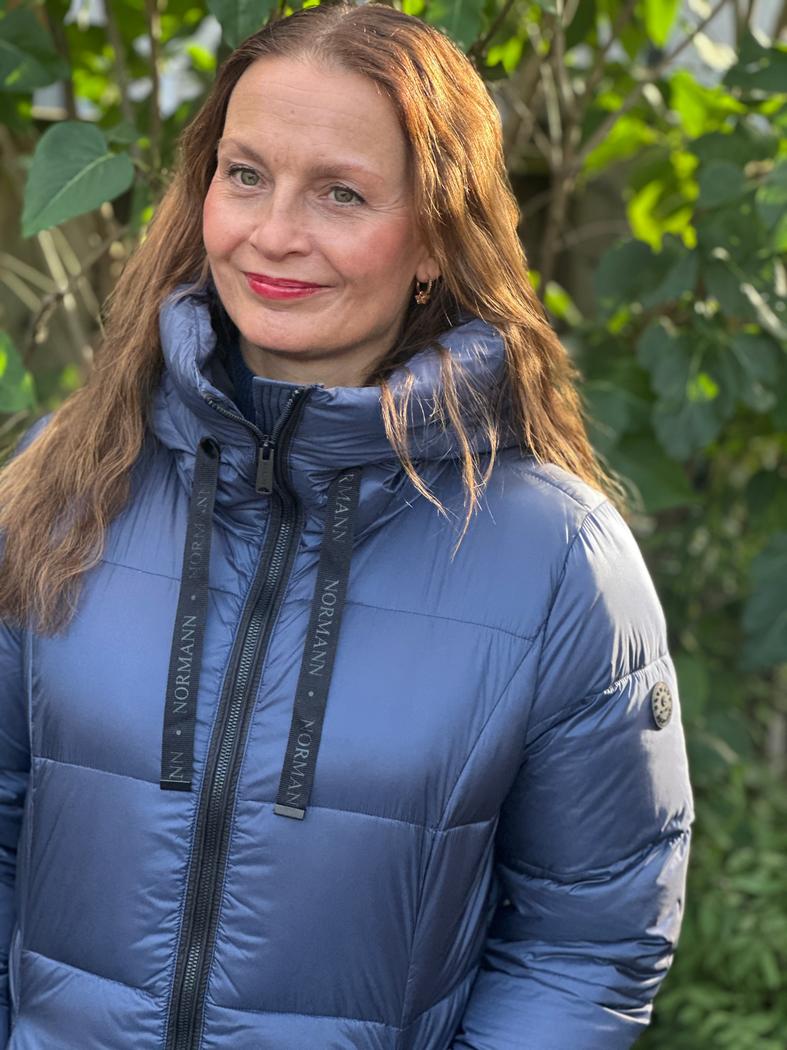 Kvinne i blå dunjakke med hette og snorer, utendørs. Jakken har synlig glidelås foran.