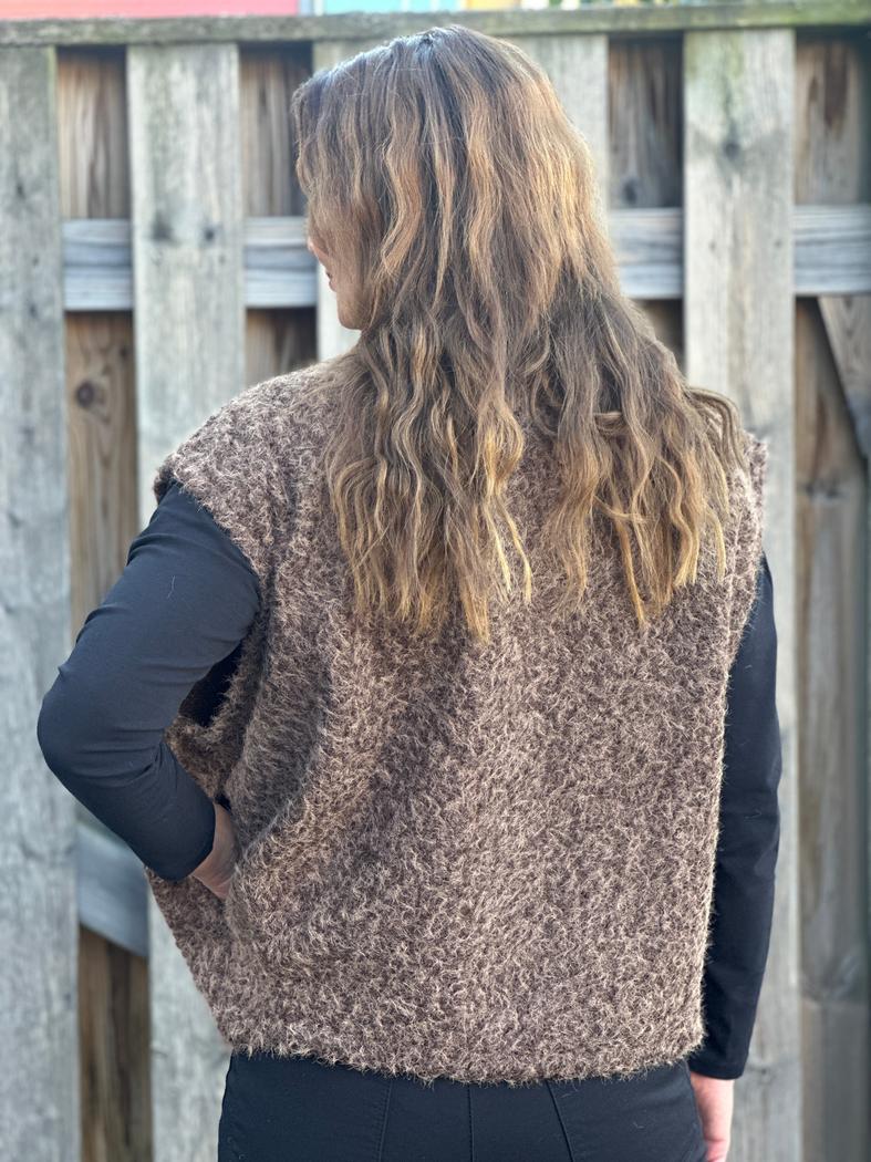 Brun strikkevest med grov tekstur, ermeløs. Personen har langt, bølgete hår. Bakgrunn av treverk.