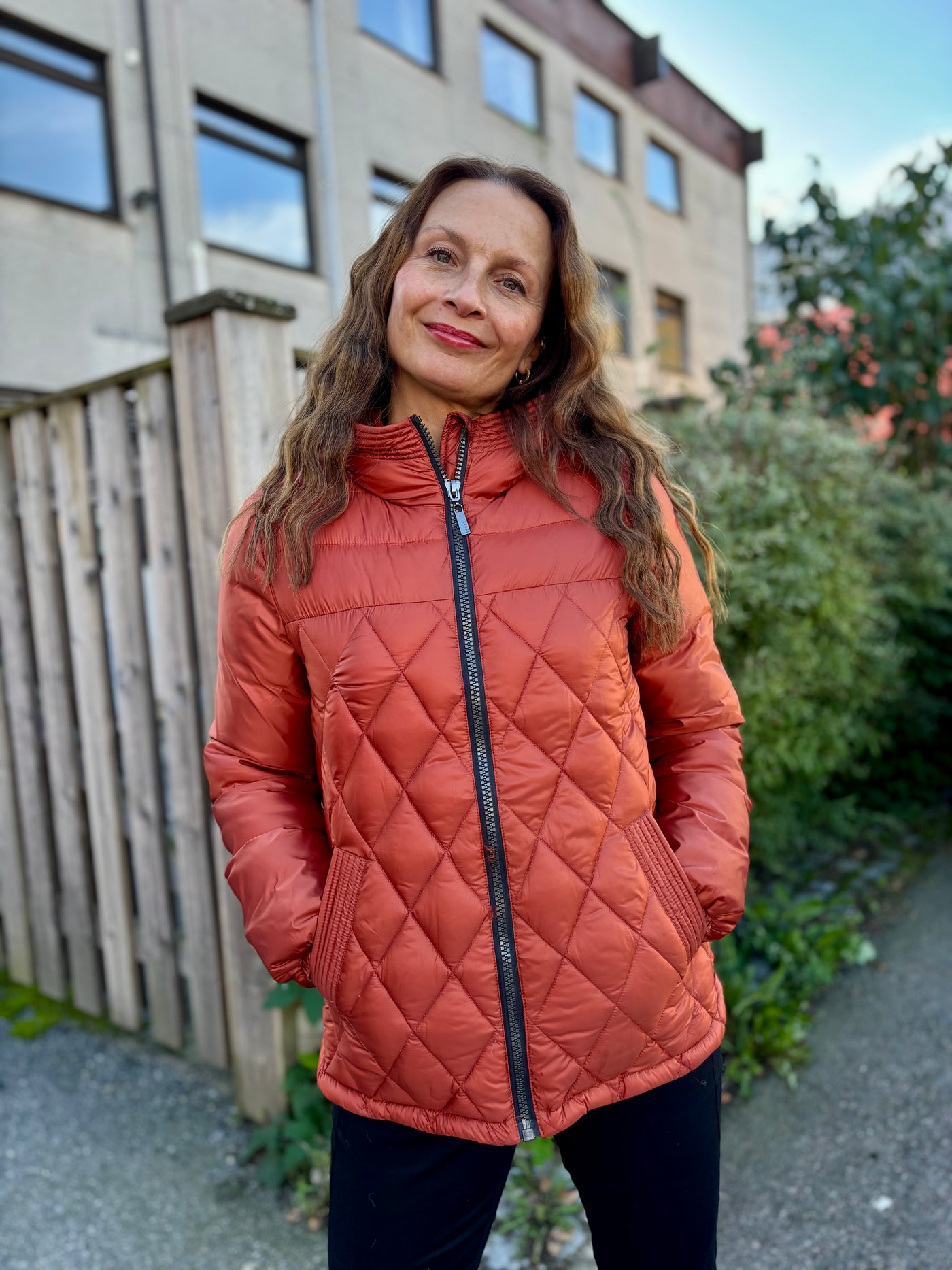 Kvinne i rød quiltet jakke med hette, stående utendørs foran et gjerde og bygning.