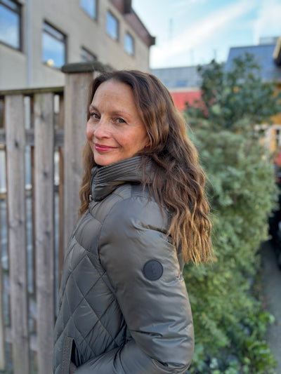 Kvinne i kort, quiltet jakke med hette. Hun står ute med bygning og grøntområde i bakgrunnen.