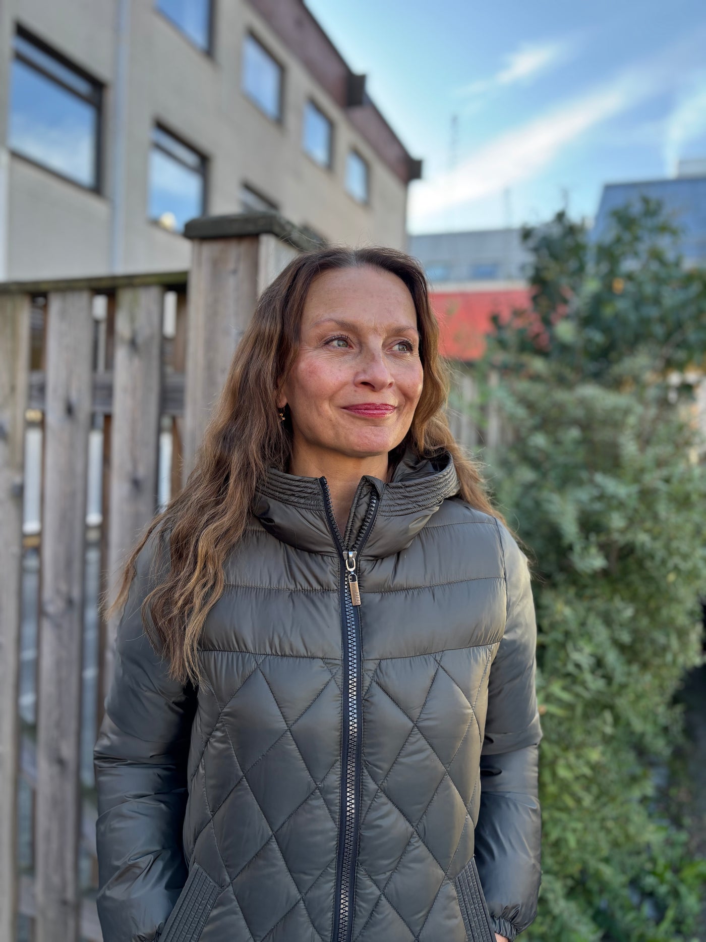 Person med mørk, quiltet jakke m/hette står foran et gjerde, med grønn bakgrunn i uskarphet.