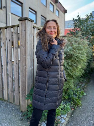 Person i lang, mørk dunjakke med avtagbar hette, foran et tregjerde og busker.