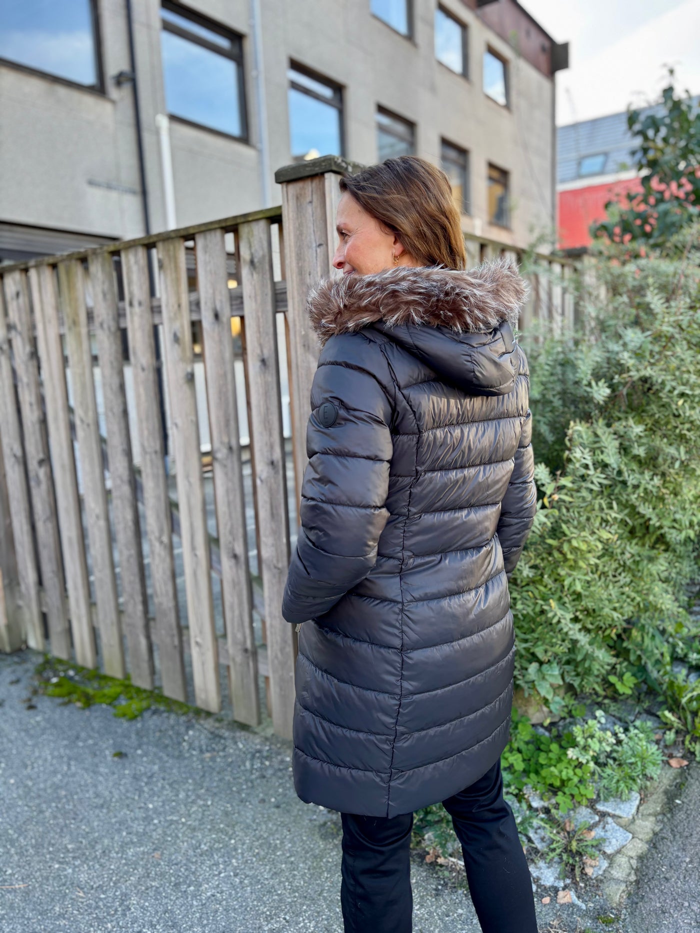Person i lang, mørk dunjakke med avtagbar hette og fuskepels, foran et gjerde og bygning.