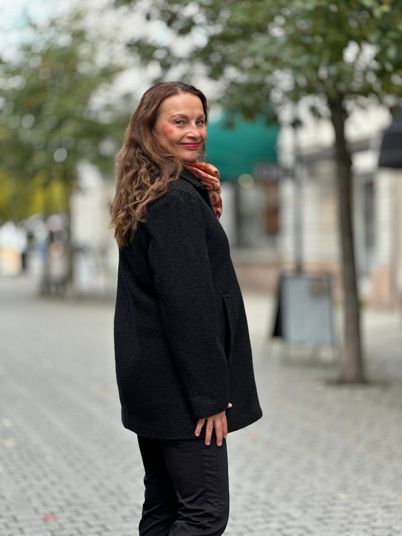 Person i svart jakke med glidelås, ute i bymiljø med trær og bygninger i bakgrunnen.