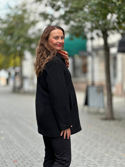 Person i svart jakke med glidelås, ute i bymiljø med trær og bygninger i bakgrunnen.