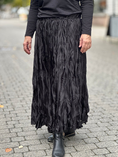 Langt, krinklet skjørt til anklene, løs passform, båret med sorte støvler på steinlagt bakgrunn.