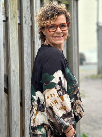 Kvinne i svart bluse med fargerikt, abstrakt mønster. Står foran et tregjerde, smiler med briller.