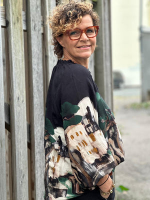 Kvinne med krøllete hår og briller i en bluse med abstrakt bygning- og treprint, smiler utendørs.