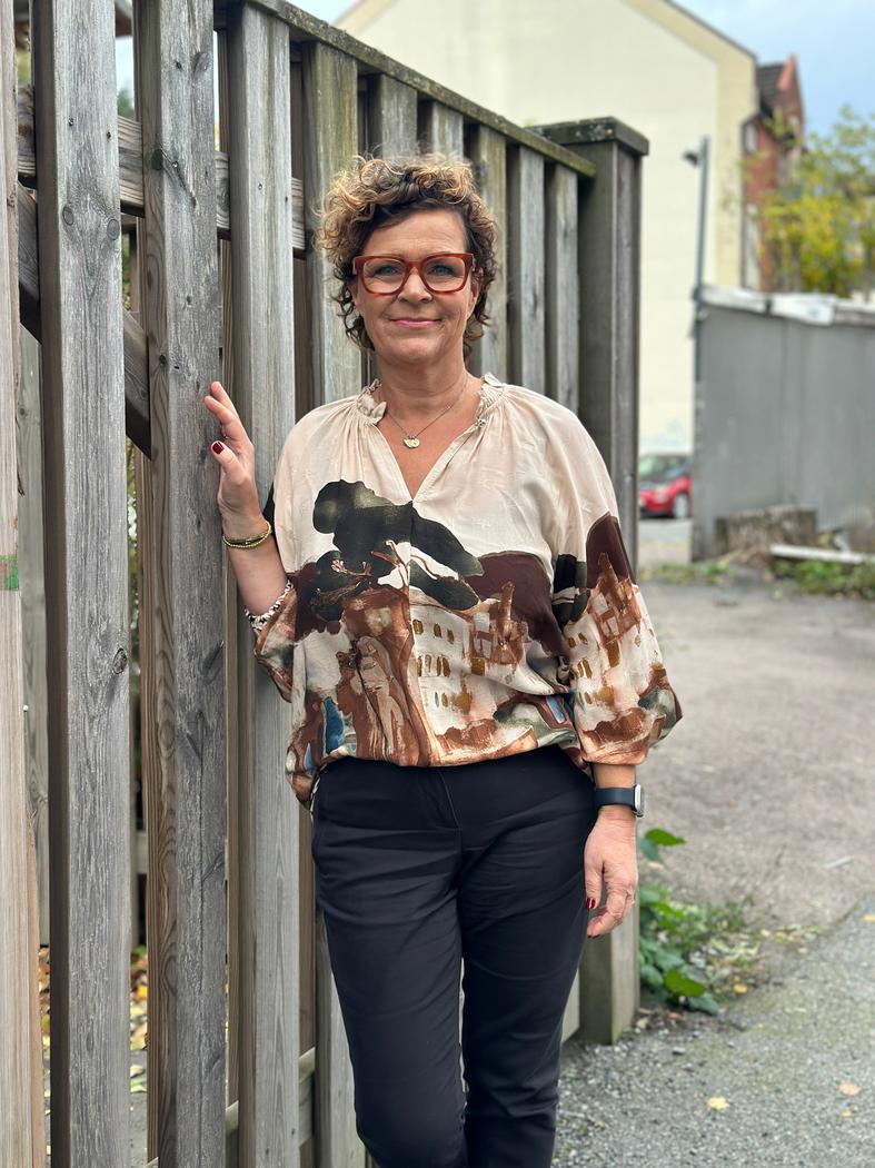 Kvinne med kort hår og briller iført viskosebluse med print, står ved et trestakitt.