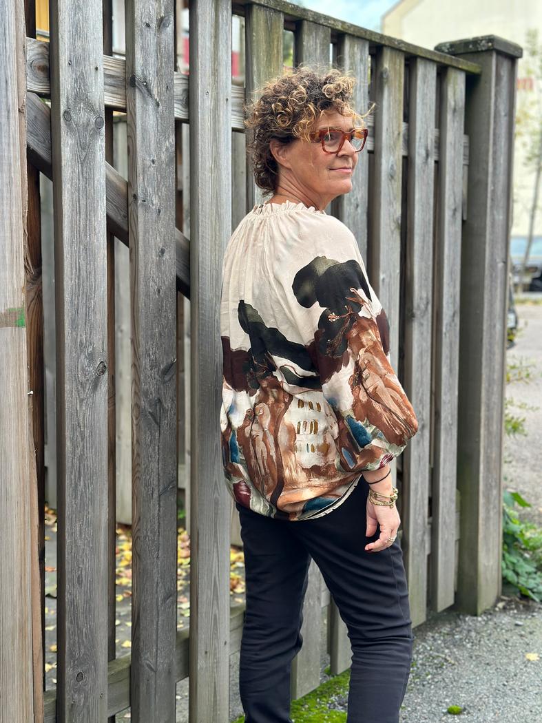 Person med krøllete hår i en fargerik viskosebluse med print, står ved et tregjerde.
