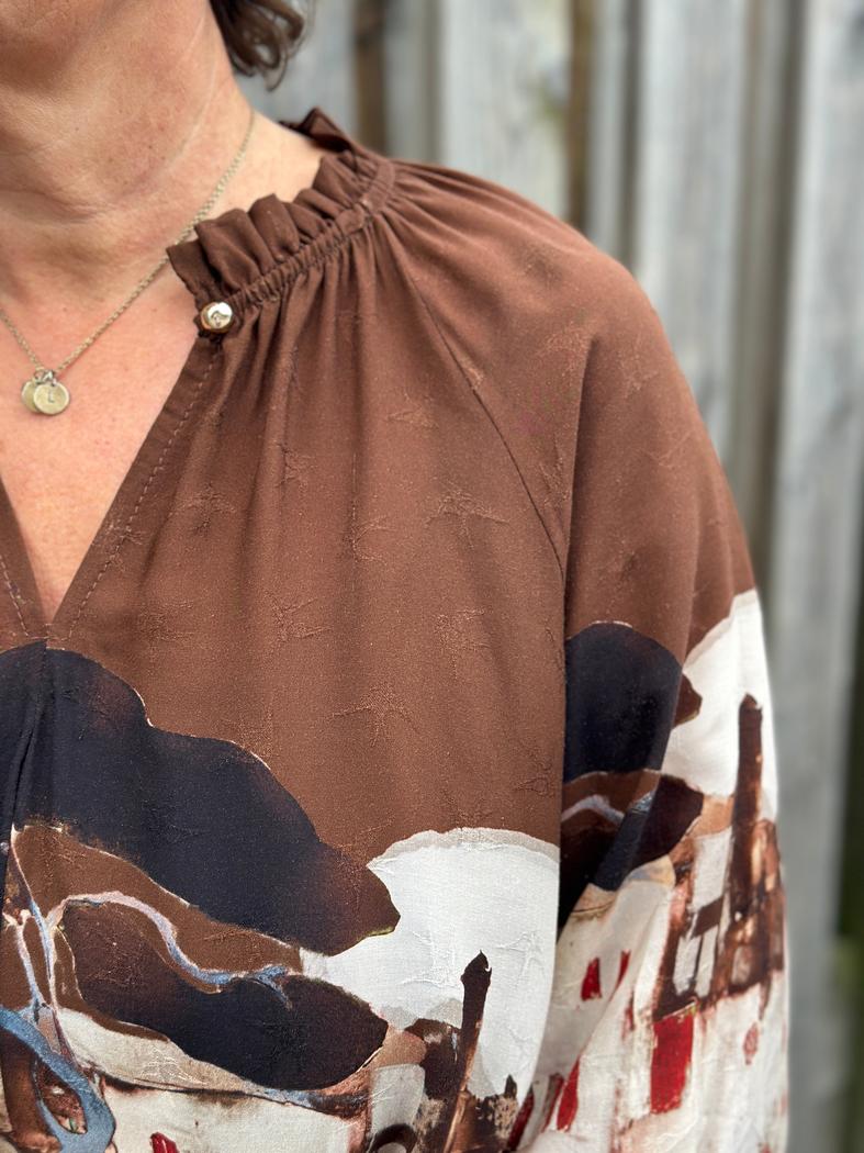 Brun bluse i viskose med rysjekrage og abstrakt mønster. Synlig knapp og del av halskjede.
