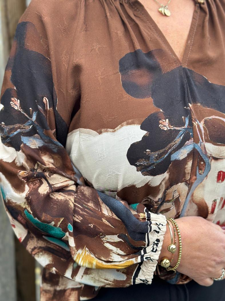 Brun bluse med abstrakt mønster og detaljerte mansjetter. Gullarmbånd på håndledd.