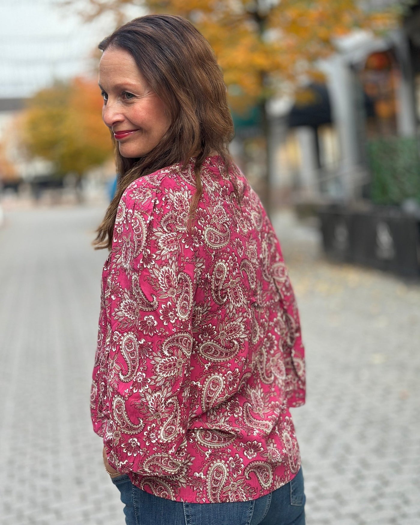 Kvinne i rosa paisleymønstret bluse med strikk på ermene, omgitt av høstfargede trær.