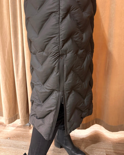 Lang, quiltet kåpe med chevronmønster, glidelås på siden. Bakgrunn: gardin og tregulv.