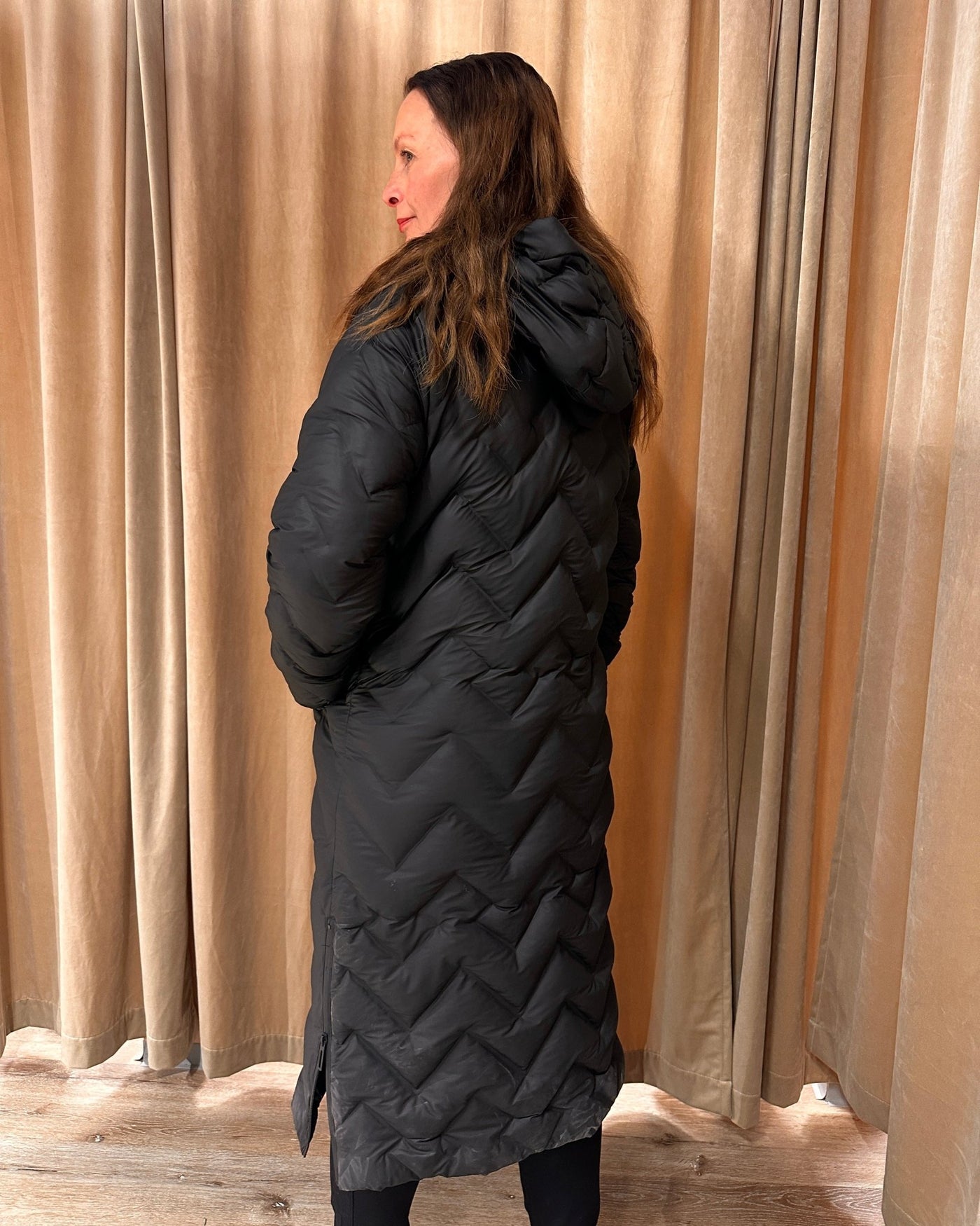 Kvinne i lang, quiltet kåpe med hette og glidelåser, beige gardiner i bakgrunnen.