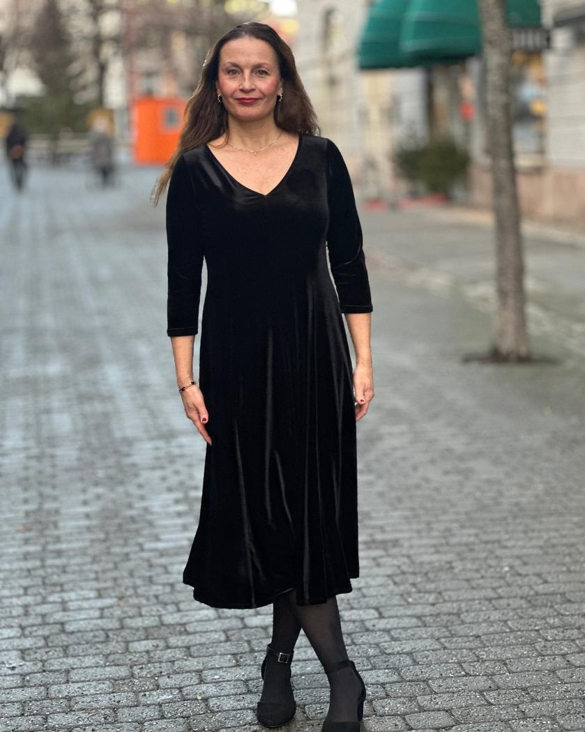 Kvinne i svart velourkjole med V-hals og trekvart ermer, står på en brosteinsgate.