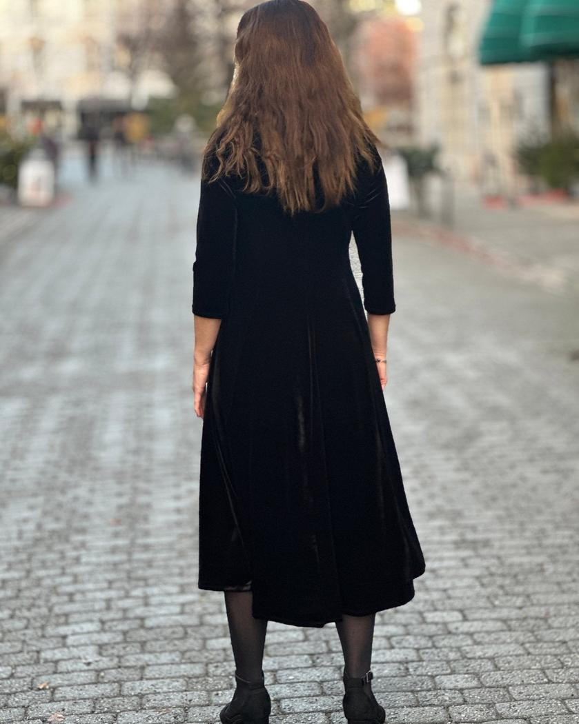 Kvinne med lang, sort velourkjole med trekvart ermer, står på brosteinsgate, sett bakfra.