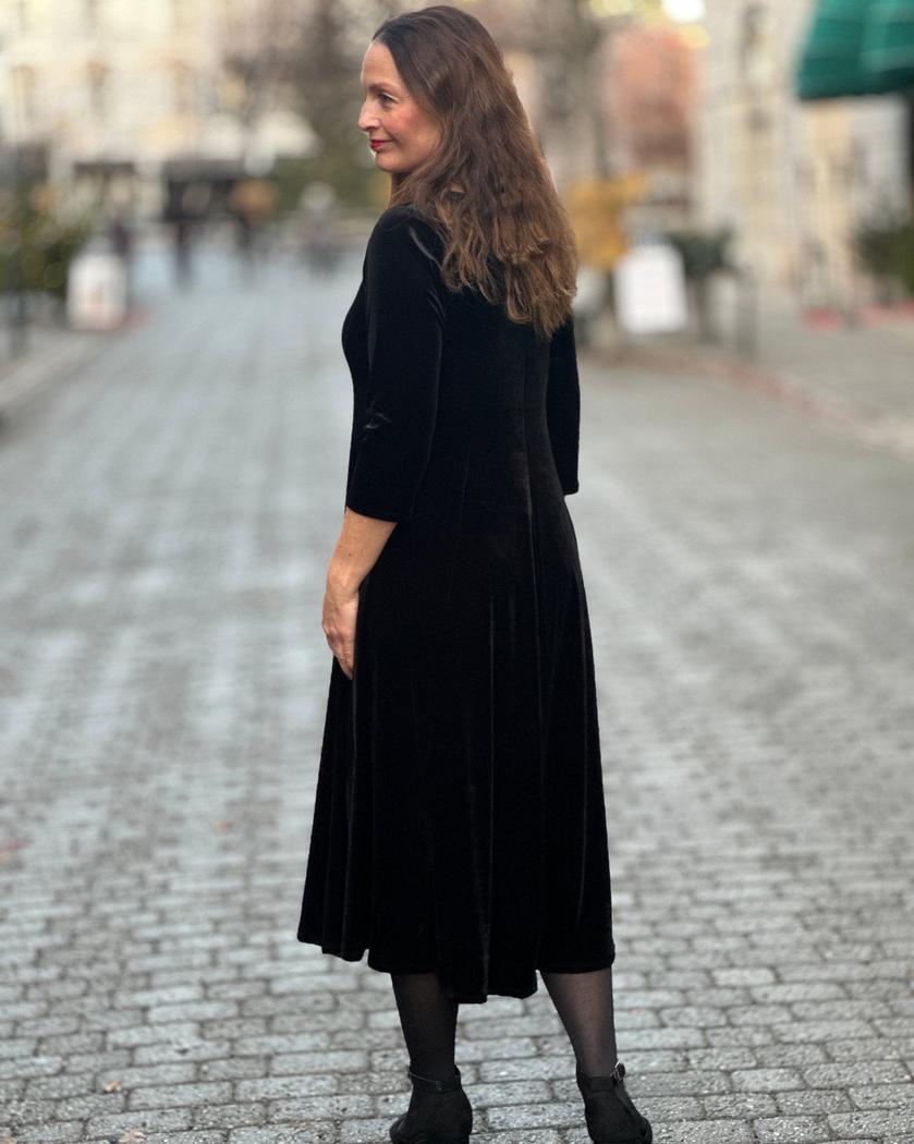 Kvinne i svart velourkjole med V-hals, trekvart ermer, midt på leggen, på brosteinsgate.