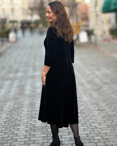Kvinne i svart velourkjole med V-hals, trekvart ermer, midt på leggen, på brosteinsgate.