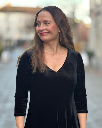 Kvinne i svart velourkjole med V-hals og trekvart ermer, smiler utendørs.