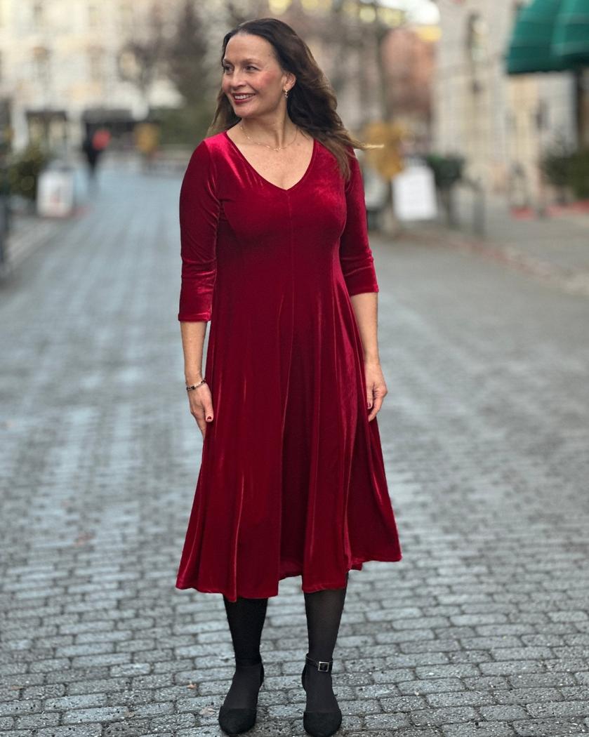 Kvinne i dyp rød velourkjole med V-hals og trekvart ermer, står på brosteinsgate.