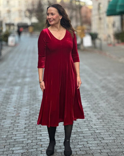 Kvinne i dyp rød velourkjole med V-hals og trekvart ermer, står på brosteinsgate.