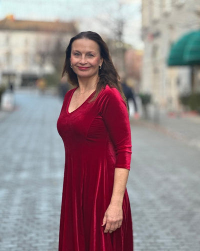 Kvinne i dyp rød velourkjole med V-hals og trekvart ermer, står på en brosteinsbelagt gate.